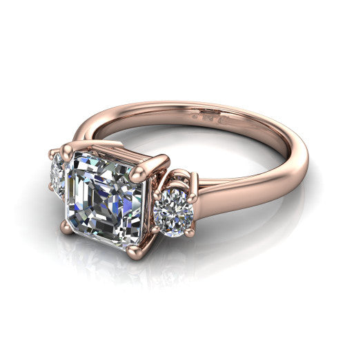 Imogen asscher three stone engagement ring in 18K Rose Gold, three-quarter perspective showing the center diamond between two round side stones on a slim polished band