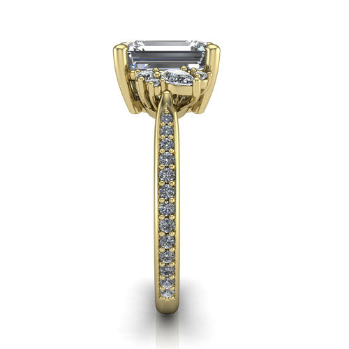 Mila emerald three stone engagement ring in 18K Yellow Gold, side profile showing the round accent cluster and the pav&eacute; diamonds running down the shank